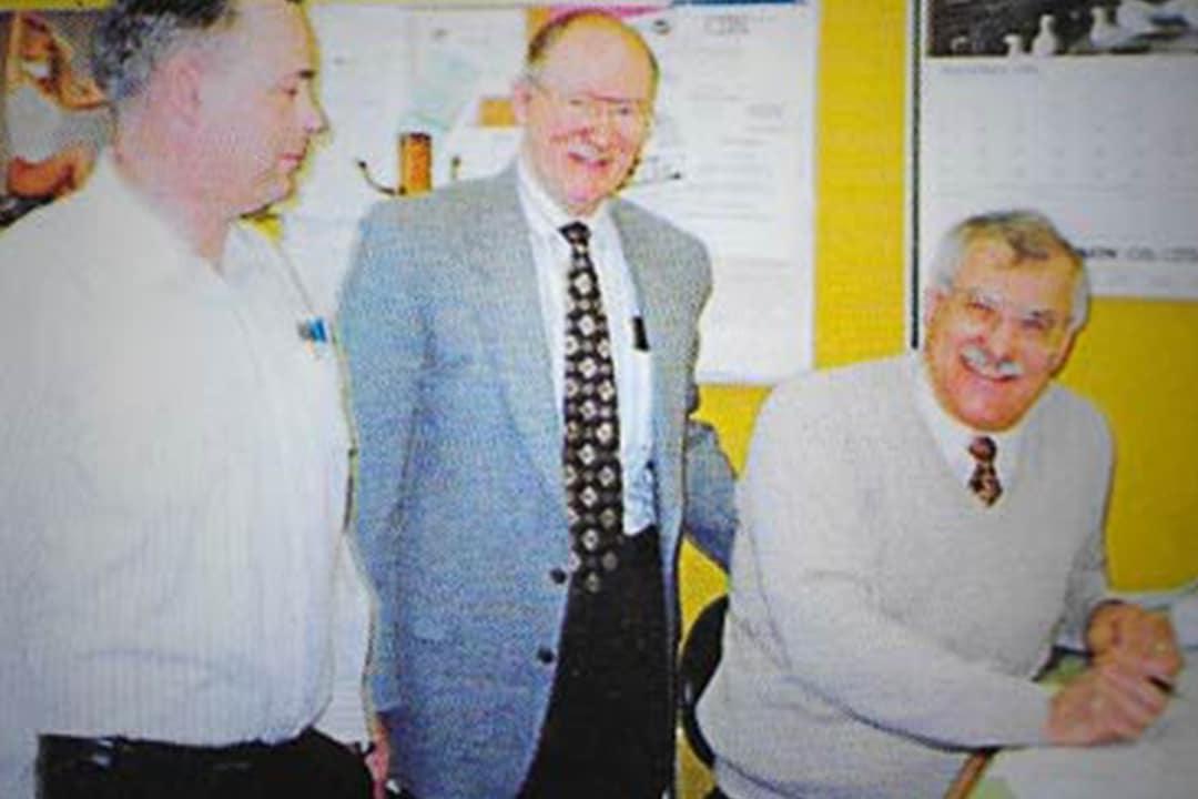 Three men in an office smiling during the late 1980s.
