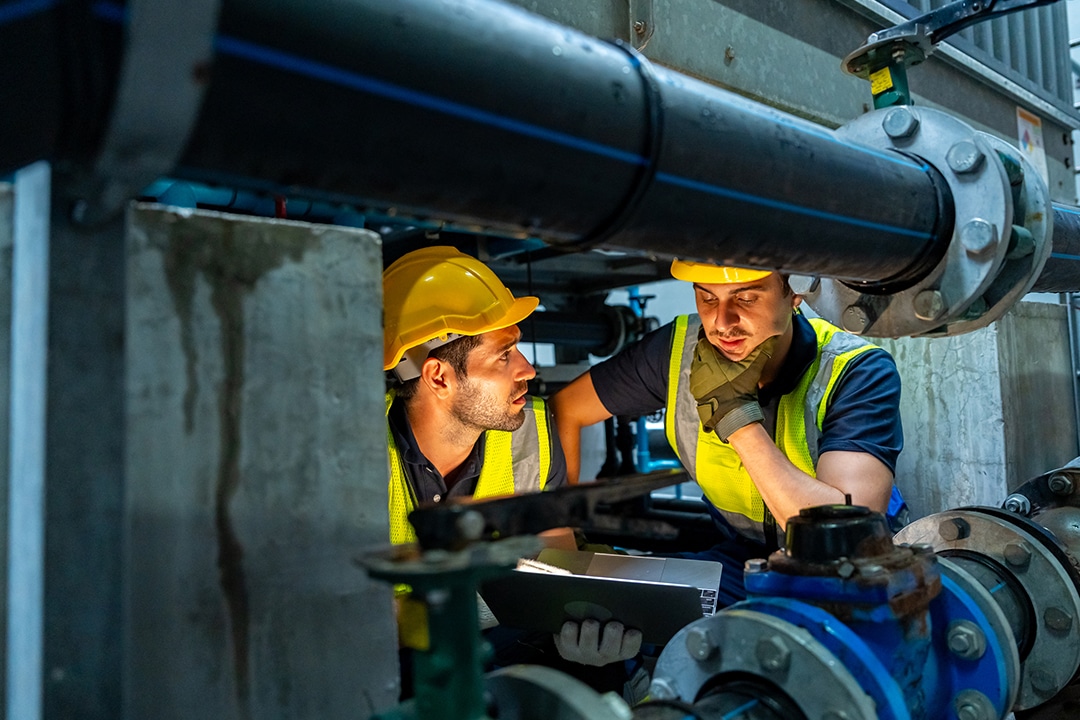 Two technicians checking a laptop under large industrial pipes.